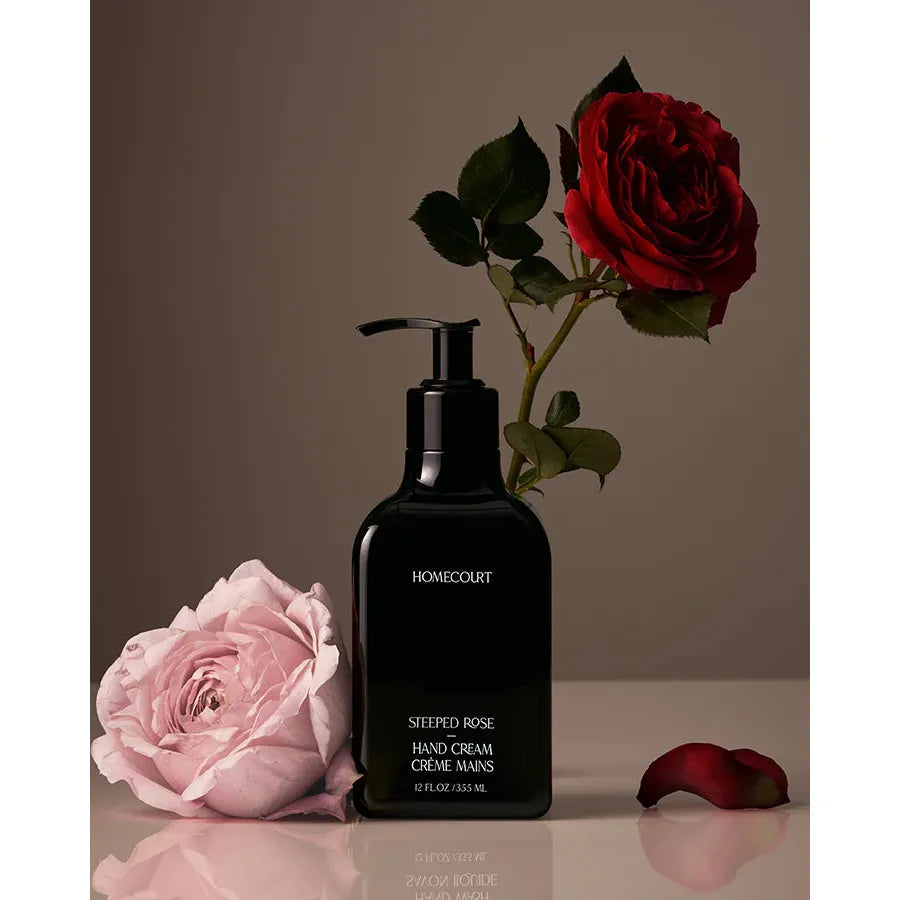 A bottle of "steeped rose hand cream" by homecourt beside a vibrant red rose and a delicate pink rose against a neutral background.