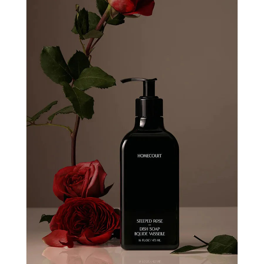 A bottle of "steeped rose dish soap" by homecourt, displayed alongside red roses against a neutral background.