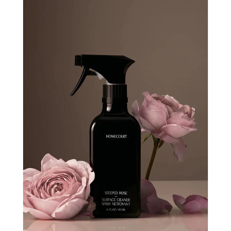 A black spray bottle of "homecourt steeped rose surface cleaner" alongside fresh pink roses against a neutral background.
