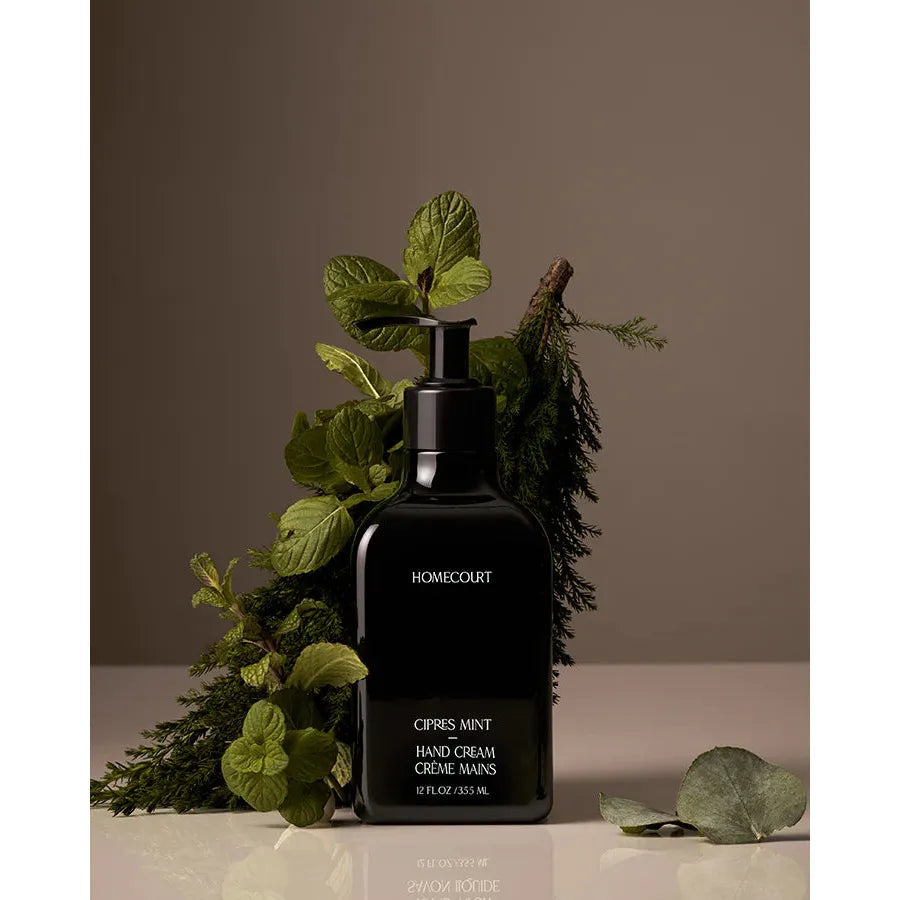 A bottle of cypress mint hand cream surrounded by green leaves and twigs against a neutral background.