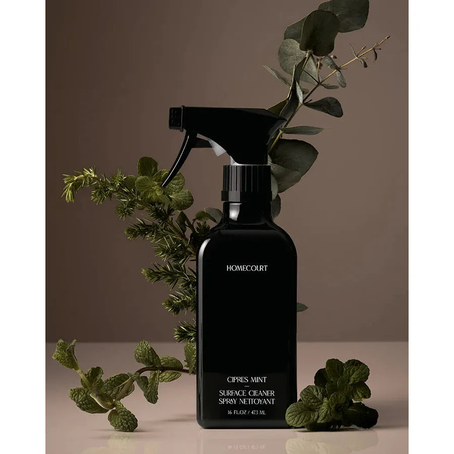 A matte black spray bottle of surface cleaner with "crisp mint" fragrance labeled "homecourt," surrounded by fresh greenery on a neutral background.