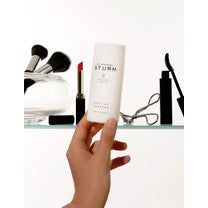 A hand reaching for a cosmetic product on a shelf with various makeup items in the background.