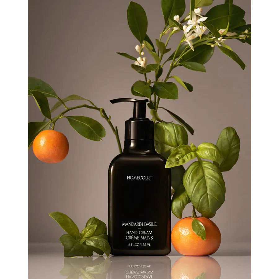 A black dispenser bottle of dermatologist tested Homecourt Hand Cream surrounded by mandarin oranges and green leaves against a beige background.