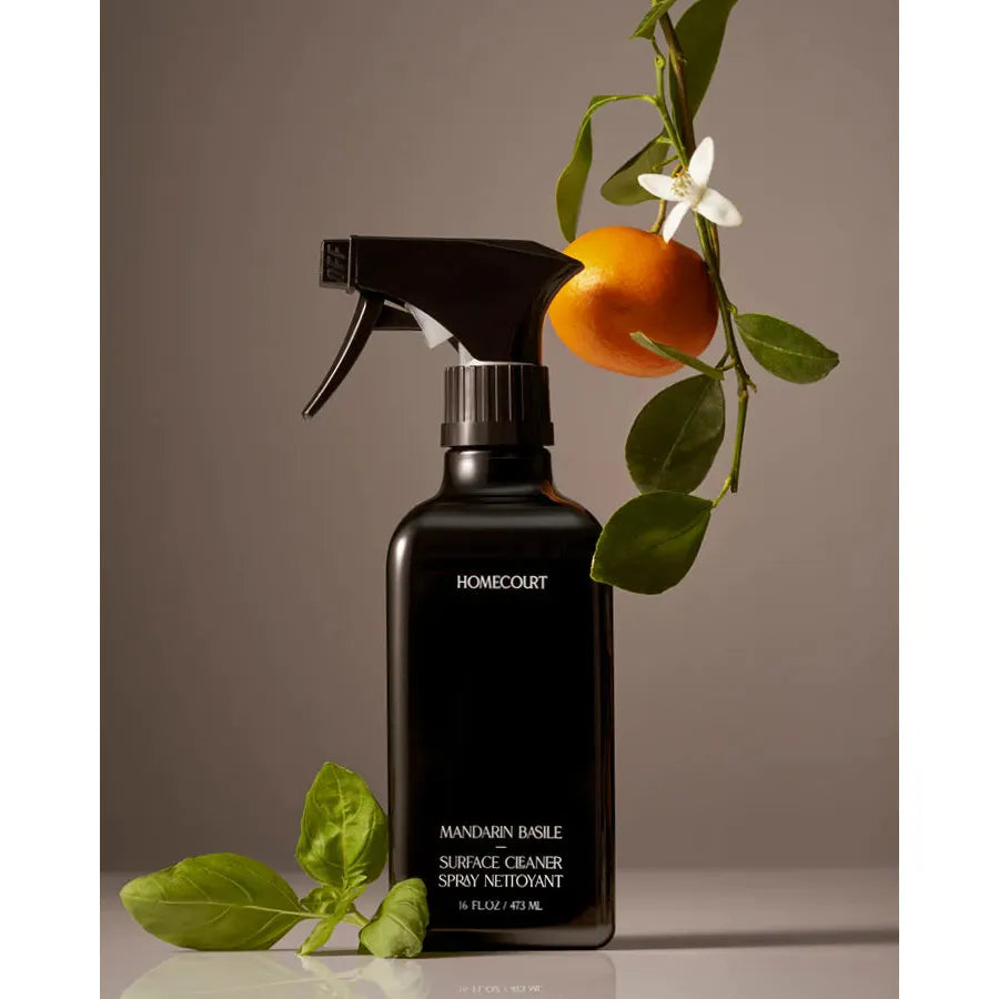 A matte black spray bottle labeled "Homecourt Surface Cleaner" by homecourt, decorated with a fresh mandarin and leaves on a neutral background, promises a streak-free finish.
