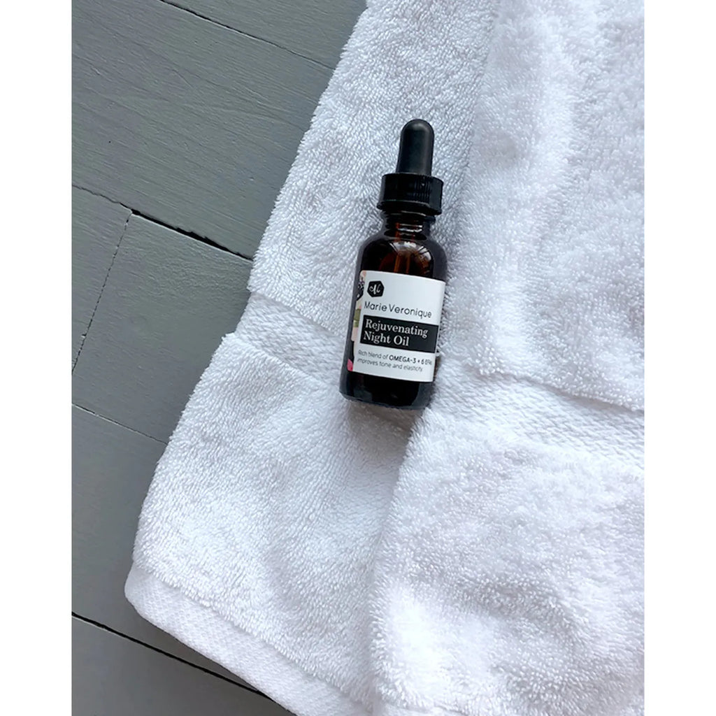A dropper bottle of rejuvenating night oil placed on a white towel with wooden surface background.