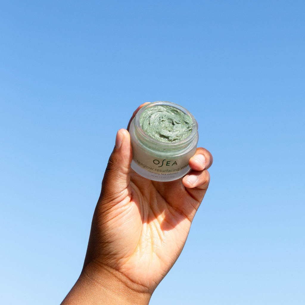 A hand holding a jar of osea skin care product against a clear blue sky.