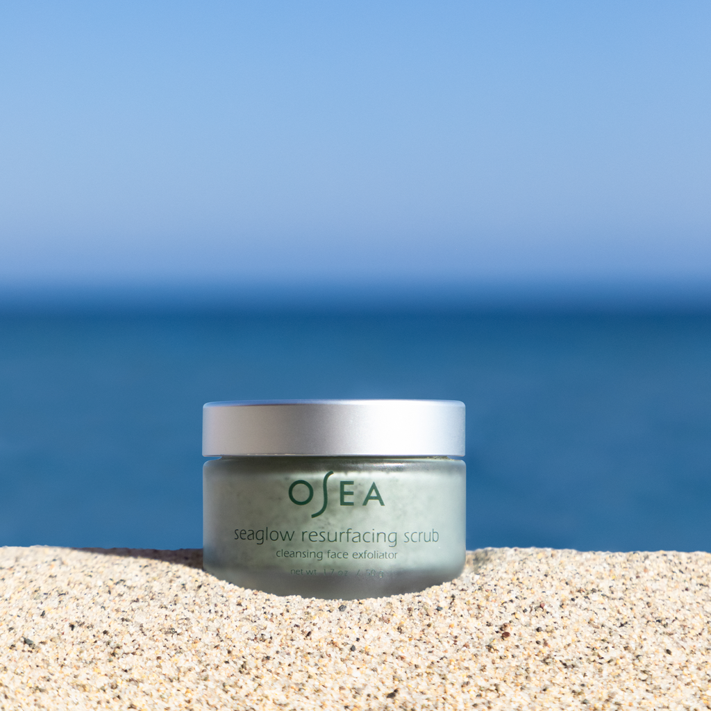 A jar of osea seaglow resurfacing scrub on a sandy surface with the ocean in the background.