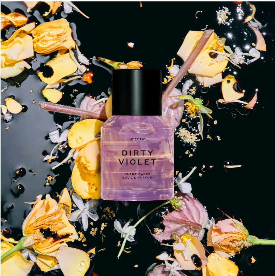 A bottle of heretic's dirty violet perfume surrounded by scattered petals and flower fragments against a dark background.