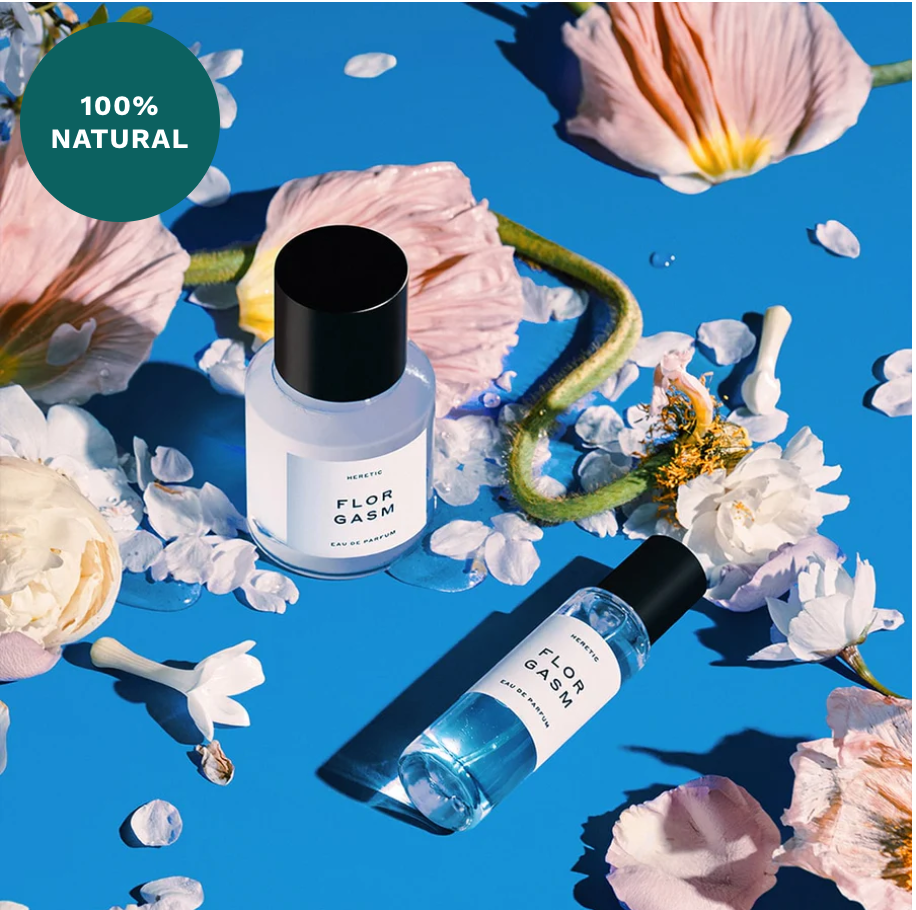 Two Heretic Parfum Florgasm bottles surrounded by scattered petals and leaves on a blue background, with a "100% natural" badge in the corner, infused with Jasmine fragrance.
