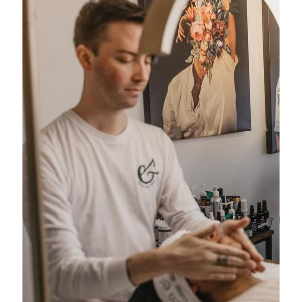 A man wearing a white shirt from Berlin Skin Matcha Mini Facials tends to products on a table in a room with a decorative floral art piece on the wall behind him.