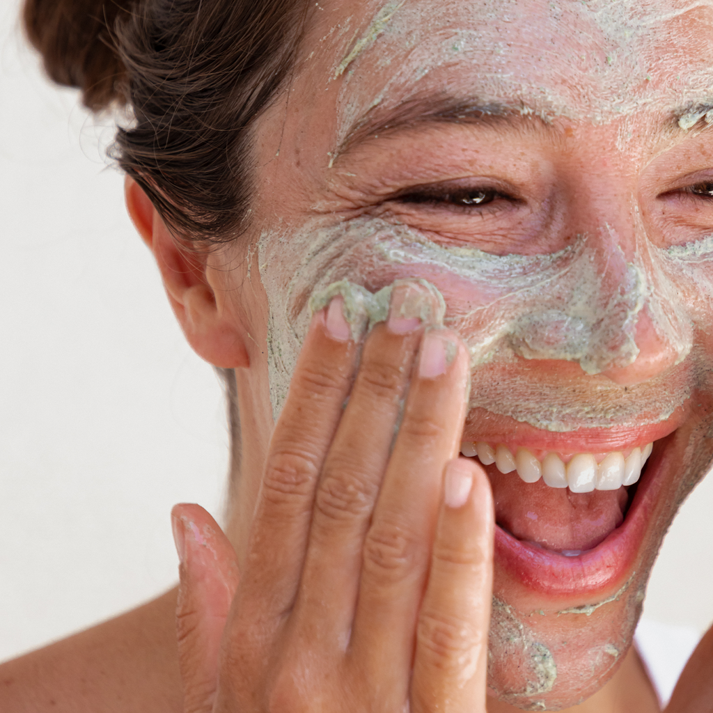 Woman applying a facial mask, smiling as she touches her face.