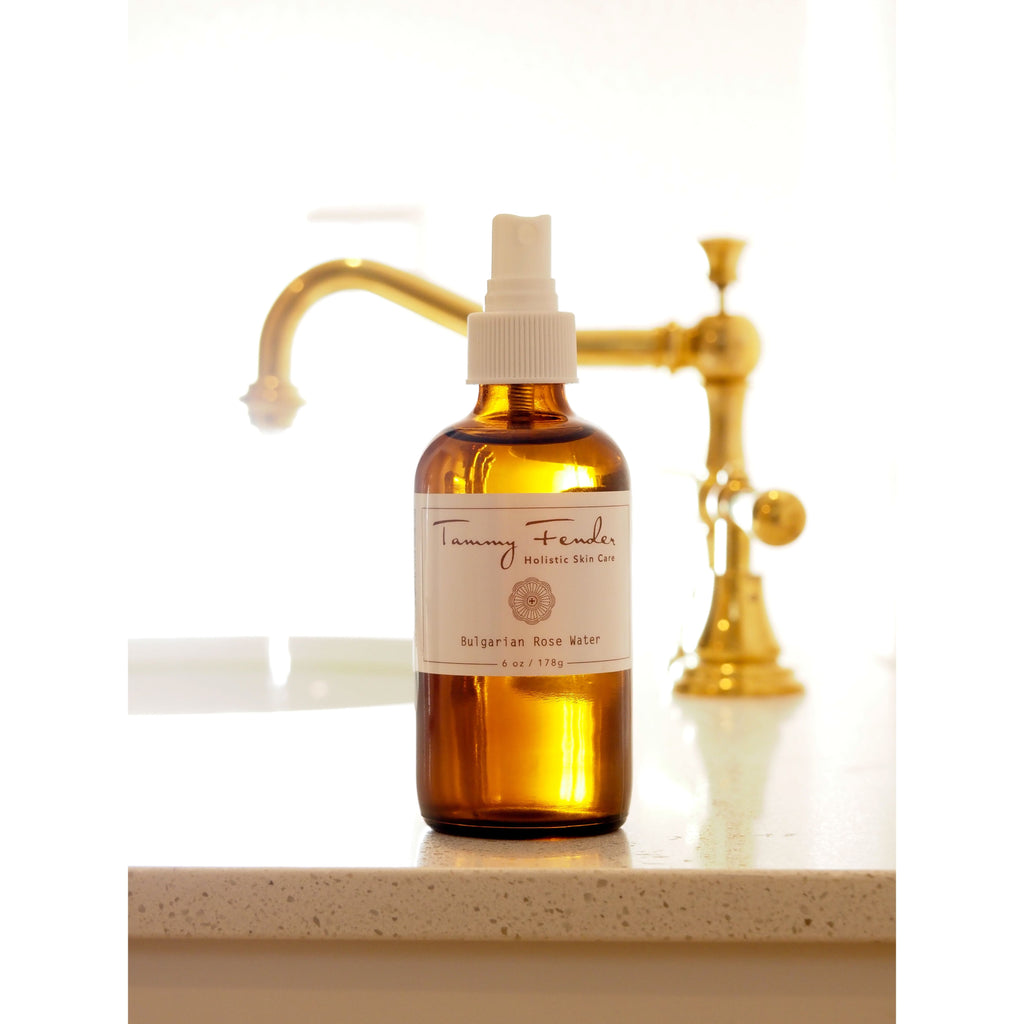 Amber glass bottle with spray nozzle labeled "tammy fender holistic skin care bulgarian rose water" placed on a surface with a gold faucet in the background.