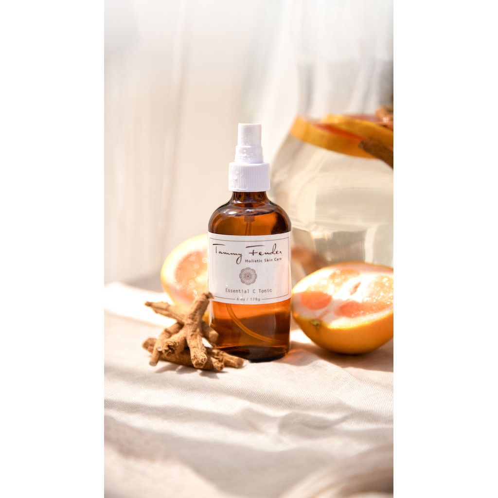 Amber glass spray bottle with a label, accompanied by sliced oranges and dried botanicals on a light fabric surface.