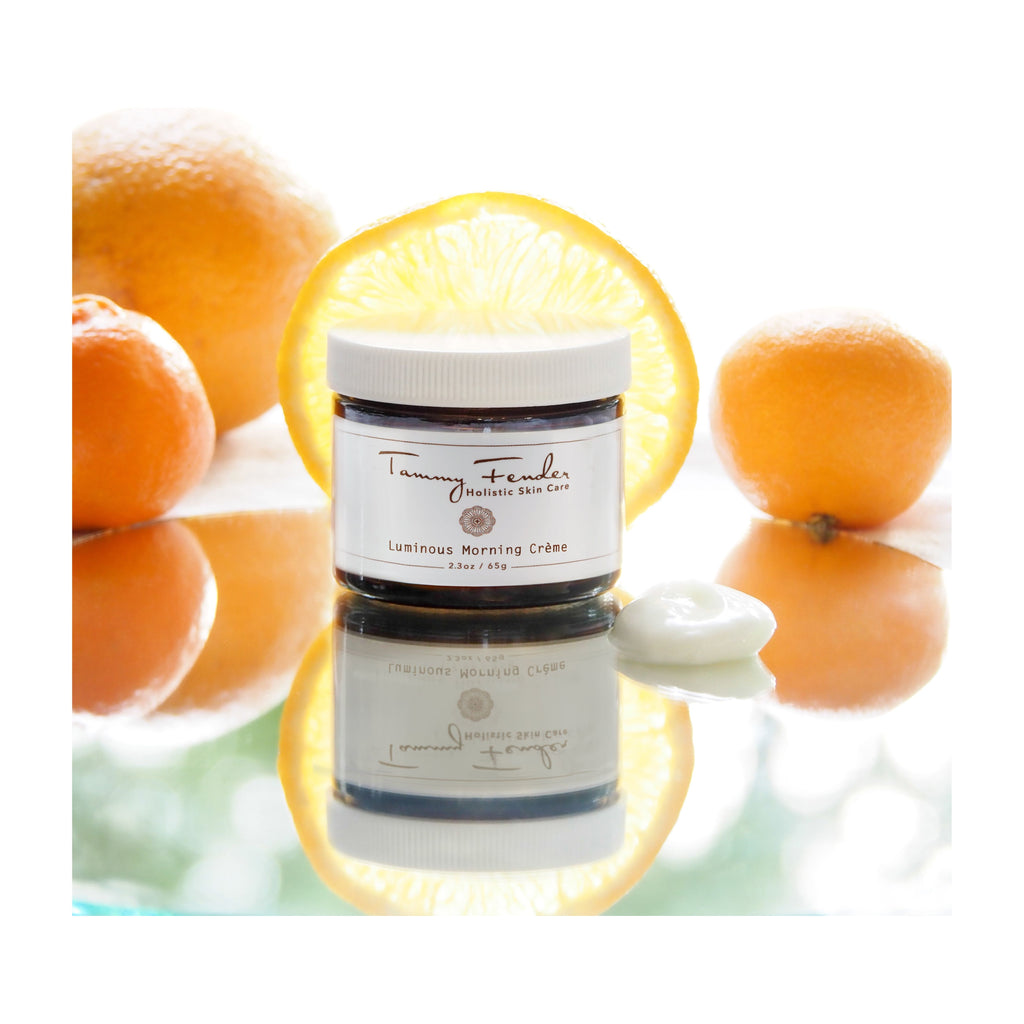 Jar of luminous morning crÃ¨me with a dollop of product on its lid, surrounded by fresh oranges and reflected on a glossy surface.