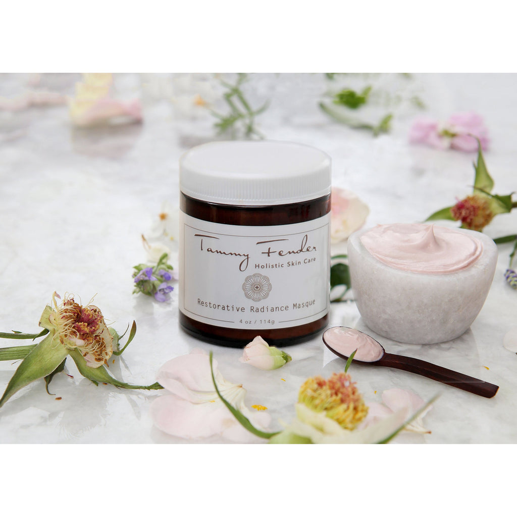 Jar of holistic skin care cream alongside a small bowl of product and a spoon, with floral petals strewn around.