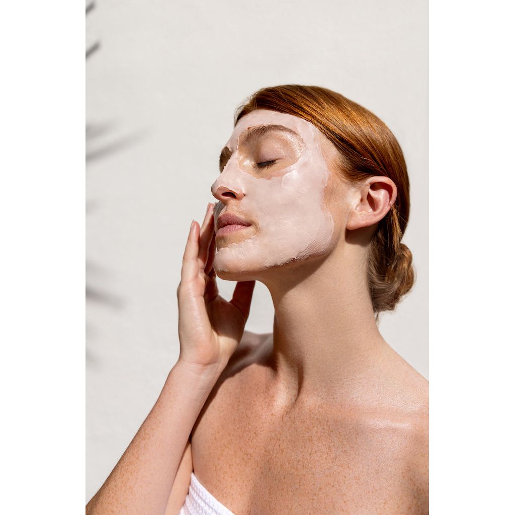 Woman applying a facial mask with eyes closed, against a light background.