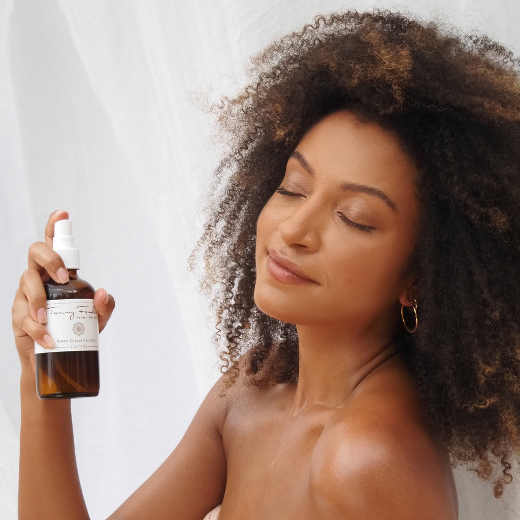 A woman with curly hair holding a spray bottle of cosmetic product with her eyes closed and a serene expression.