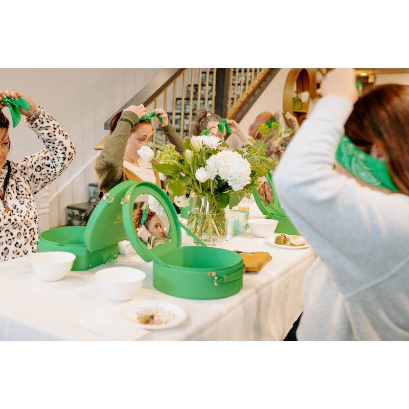 Women trying on green headbands in front of a mirror at a table with food and flowers.