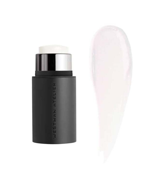 The Westman Atelie Mini Petite Lit Up Cream Highlighter Stick, in a black container with a white cosmetic product, sits next to a smear showcasing its gel highlighter. Set against a white background, it promises radiant clarity and a glassy finish.