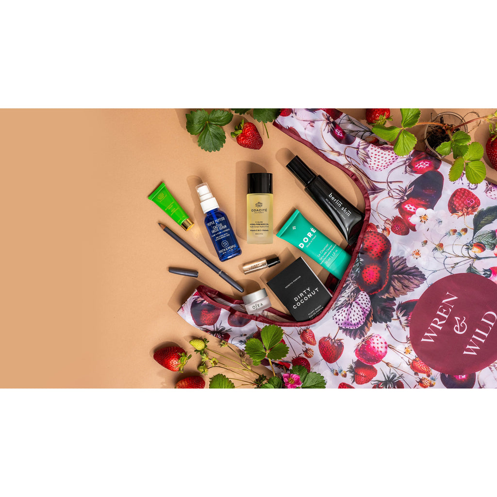 A variety of Berry Beautiful: 9-Piece Mother's Day Gift for Plant-Based Beauty Lovers skincare products and makeup items displayed on a floral fabric with strawberries scattered around.