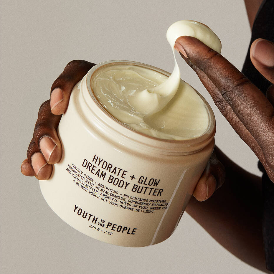 A person applying a cream enriched with niacinamide from a large jar labeled "Youth To The People Superberry Firm + Glow Dream Body Butter".