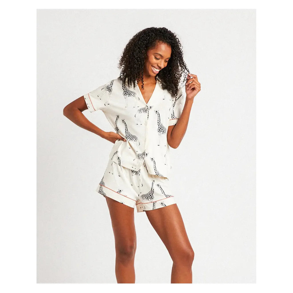 A person with curly hair wears a charming Chelsea Peers Organic Cotton Giraffe Print pajama set, smiling and looking down to their right in a bright setting.
