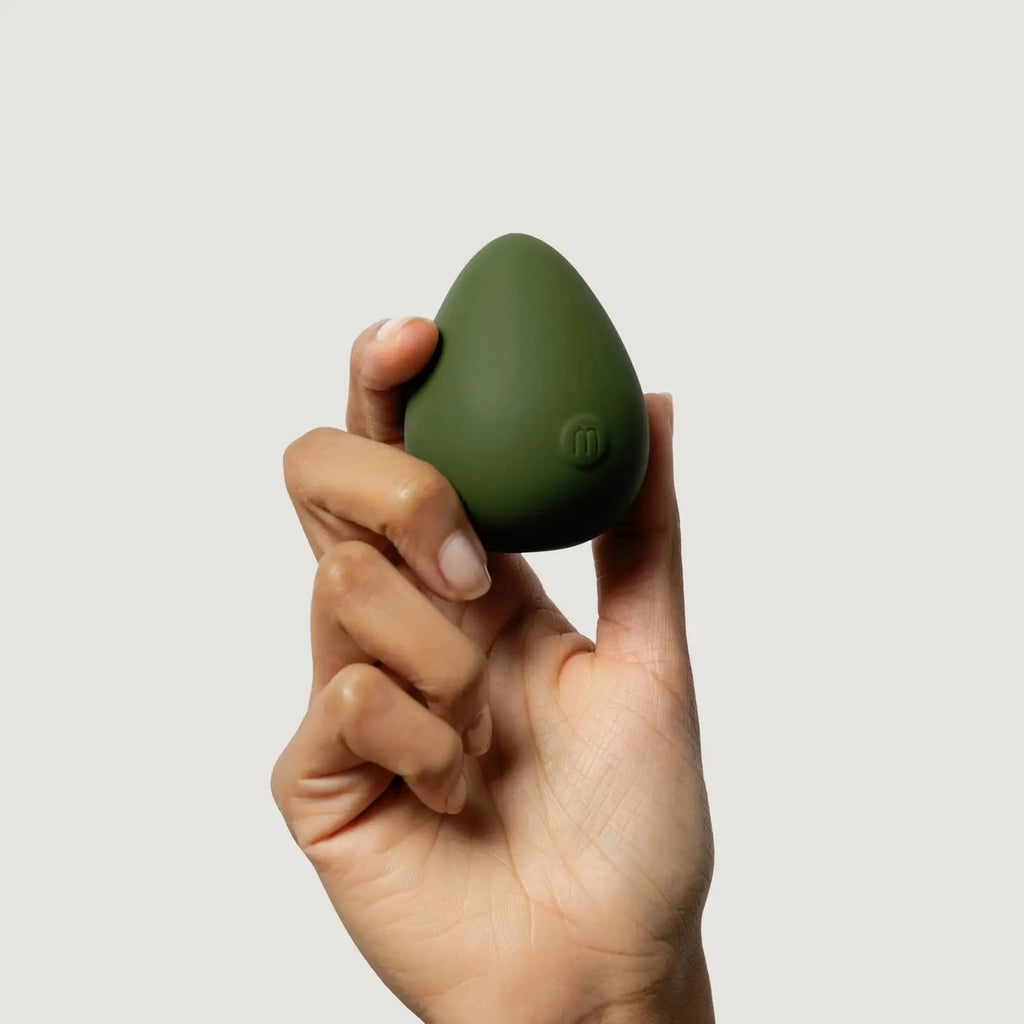 A hand holding a green, egg-shaped beauty blender against a neutral background.