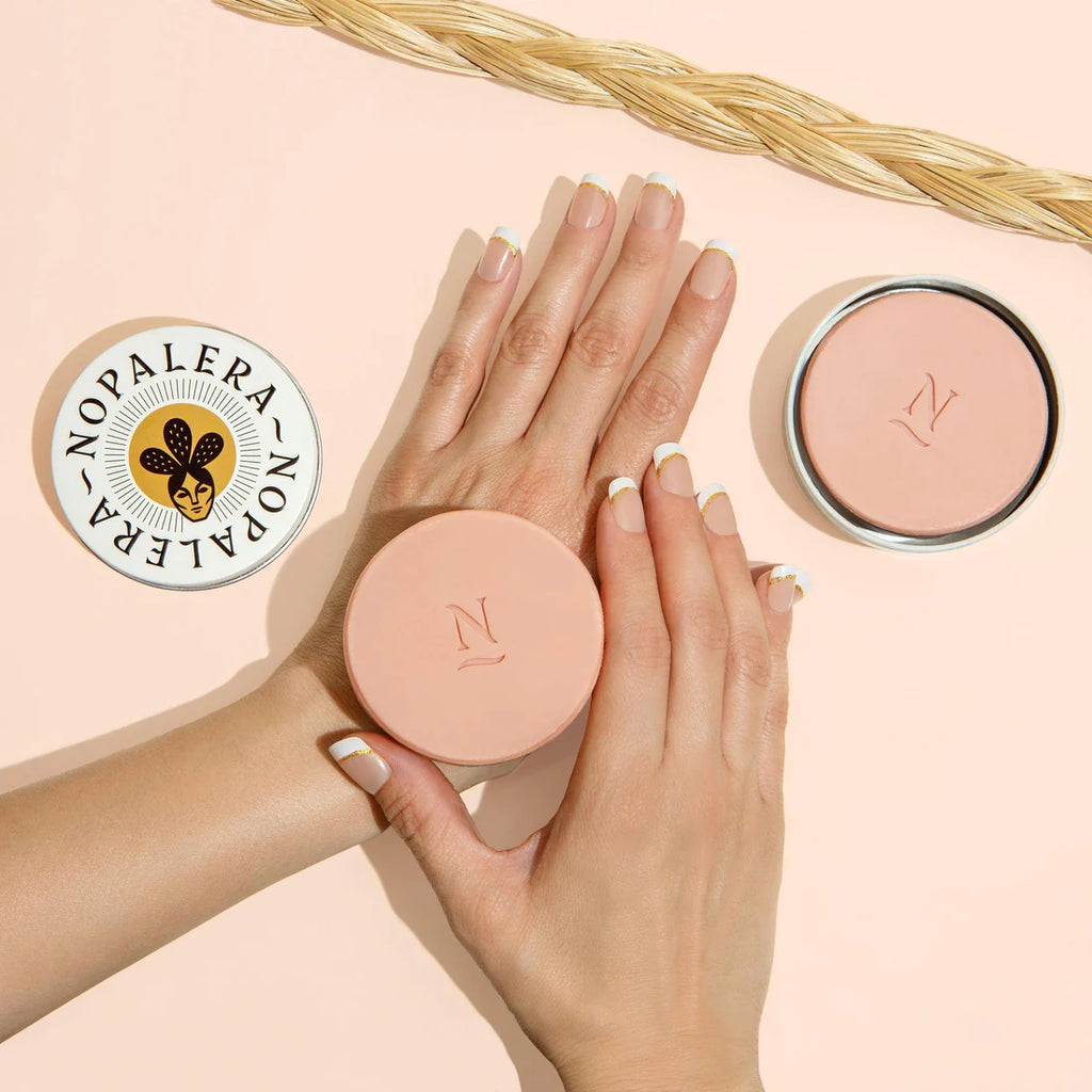 A person showing off their manicured hands with a neutral nail color next to two cosmetic jars on a peach-colored background.