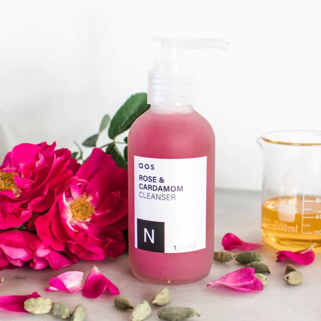 Bottle of rose and cardamom cleanser with fresh roses and dried botanicals on a neutral background.