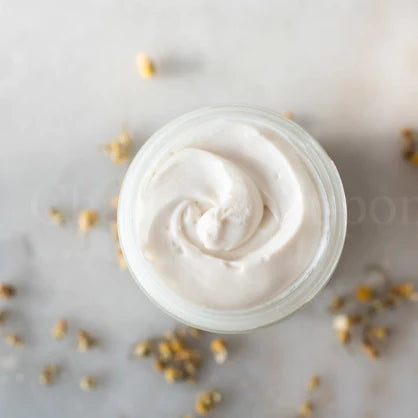 Open jar of creamy skincare product with scattered dry flowers around on a marble surface.