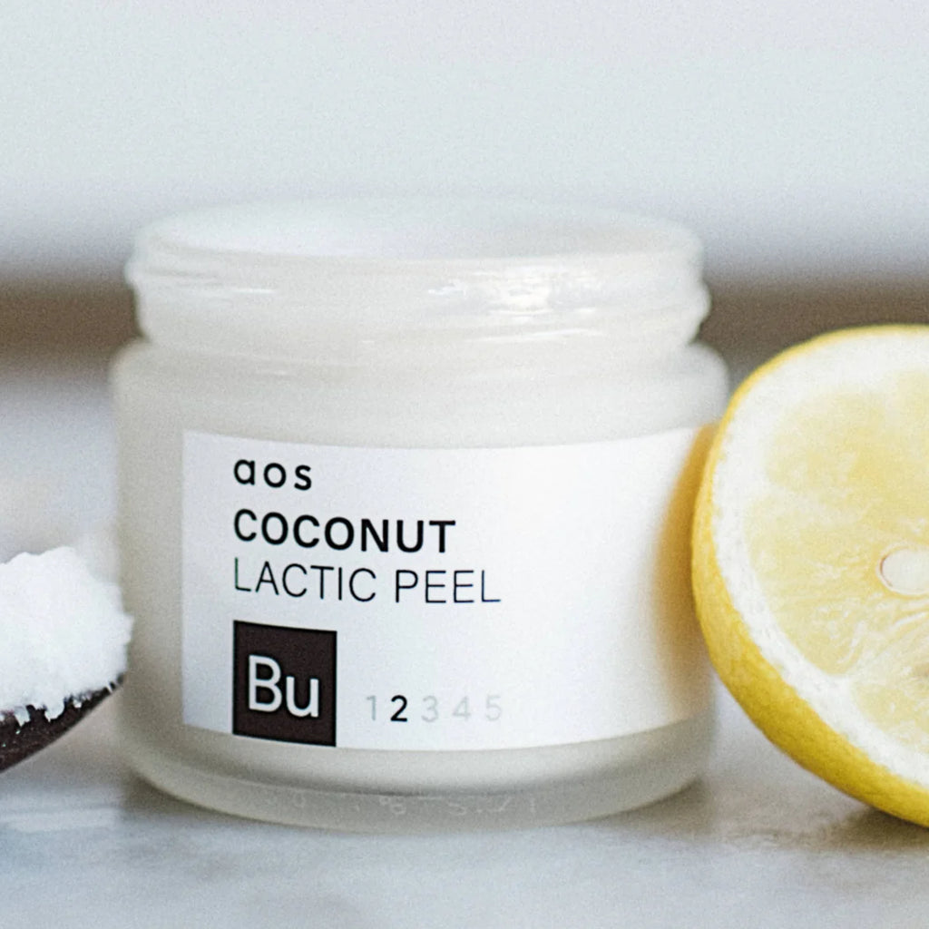 A jar of coconut lactic peel product with a slice of lemon and a spoonful of coconut flakes beside it.