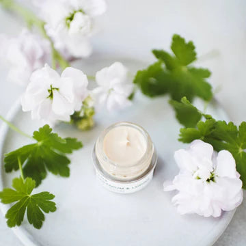 A jar of cream surrounded by white flowers and green leaves on a light-colored surface.