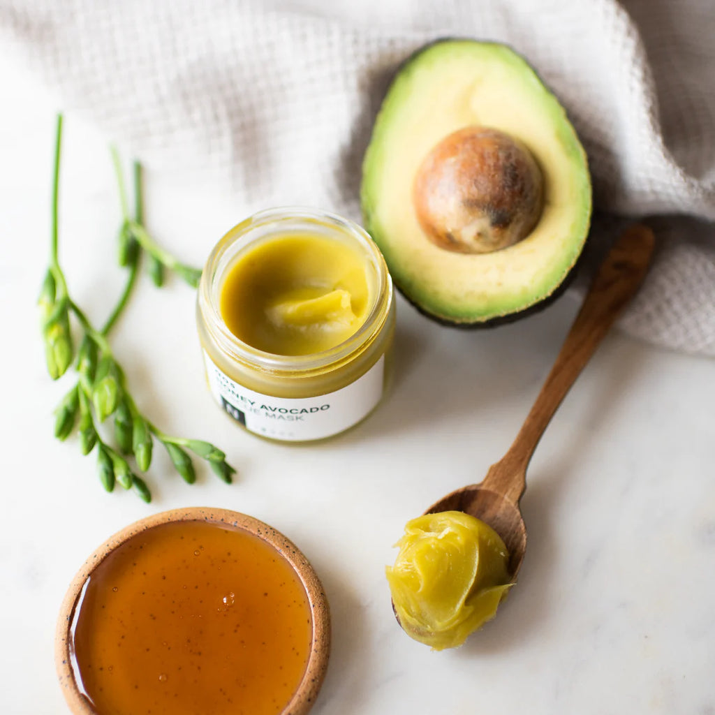 A jar of avocado spread, a wooden spoon with a dollop of spread, half an avocado, and a small bowl of dressing on a marble surface.