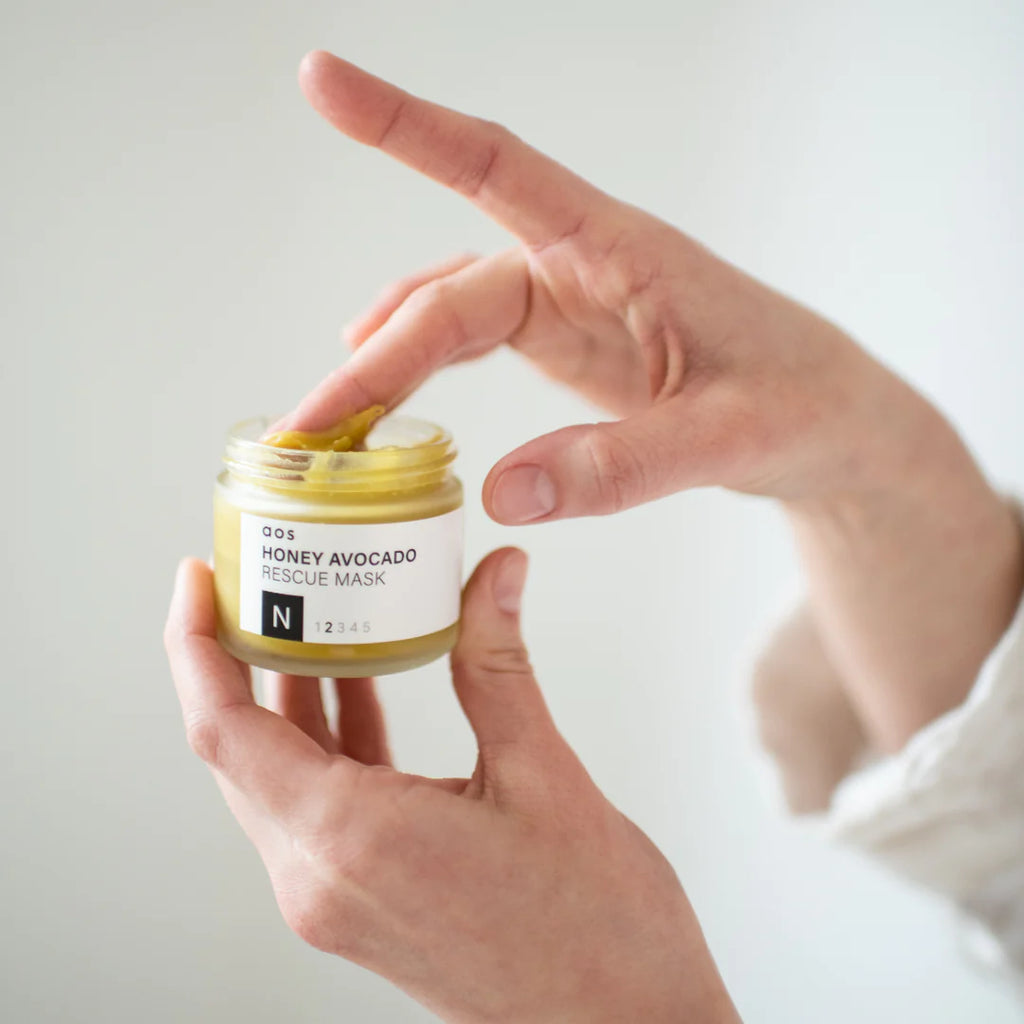 Person holding a small jar of honey avocado rescue mask and dipping a finger into it.