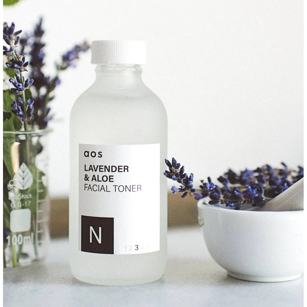 A bottle of lavender and aloe facial toner on a countertop, with lavender stems and a bowl of lavender blooms in the background.
