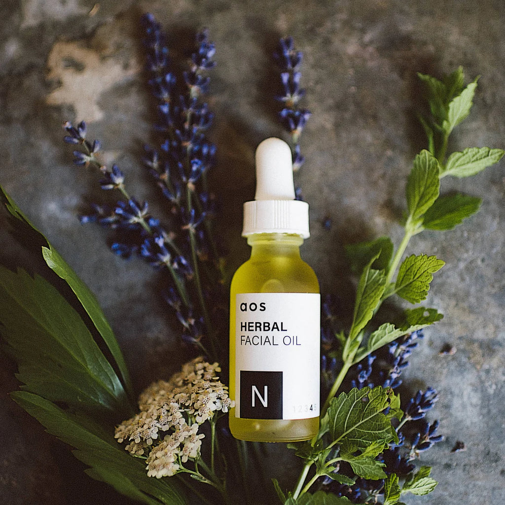 Bottle of herbal facial oil surrounded by fresh plants and flowers on a stone surface.