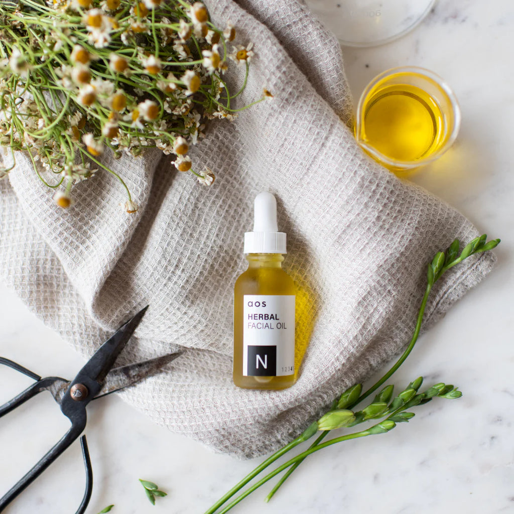 A bottle of herbal facial oil with sprigs of greenery and a pair of scissors on a textured cloth.