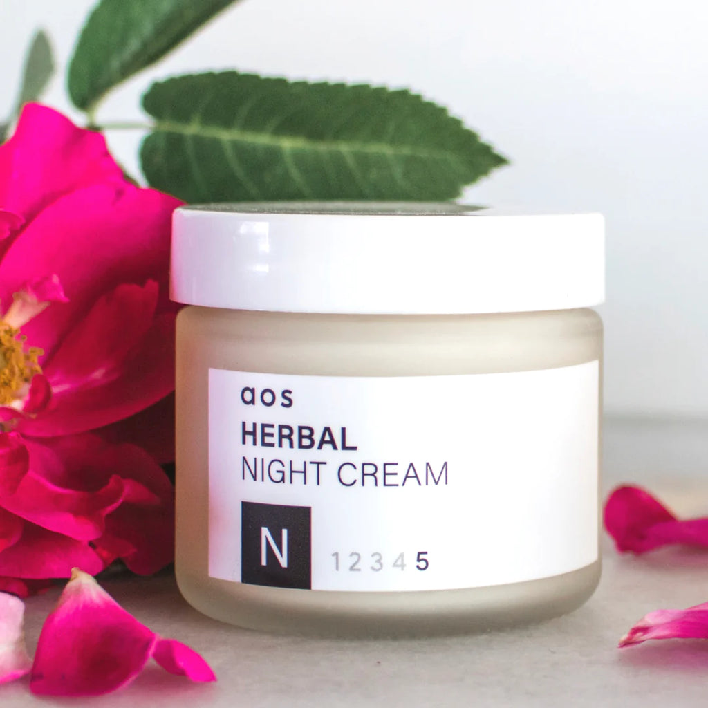 A jar of herbal night cream surrounded by vibrant pink flowers and green leaves.