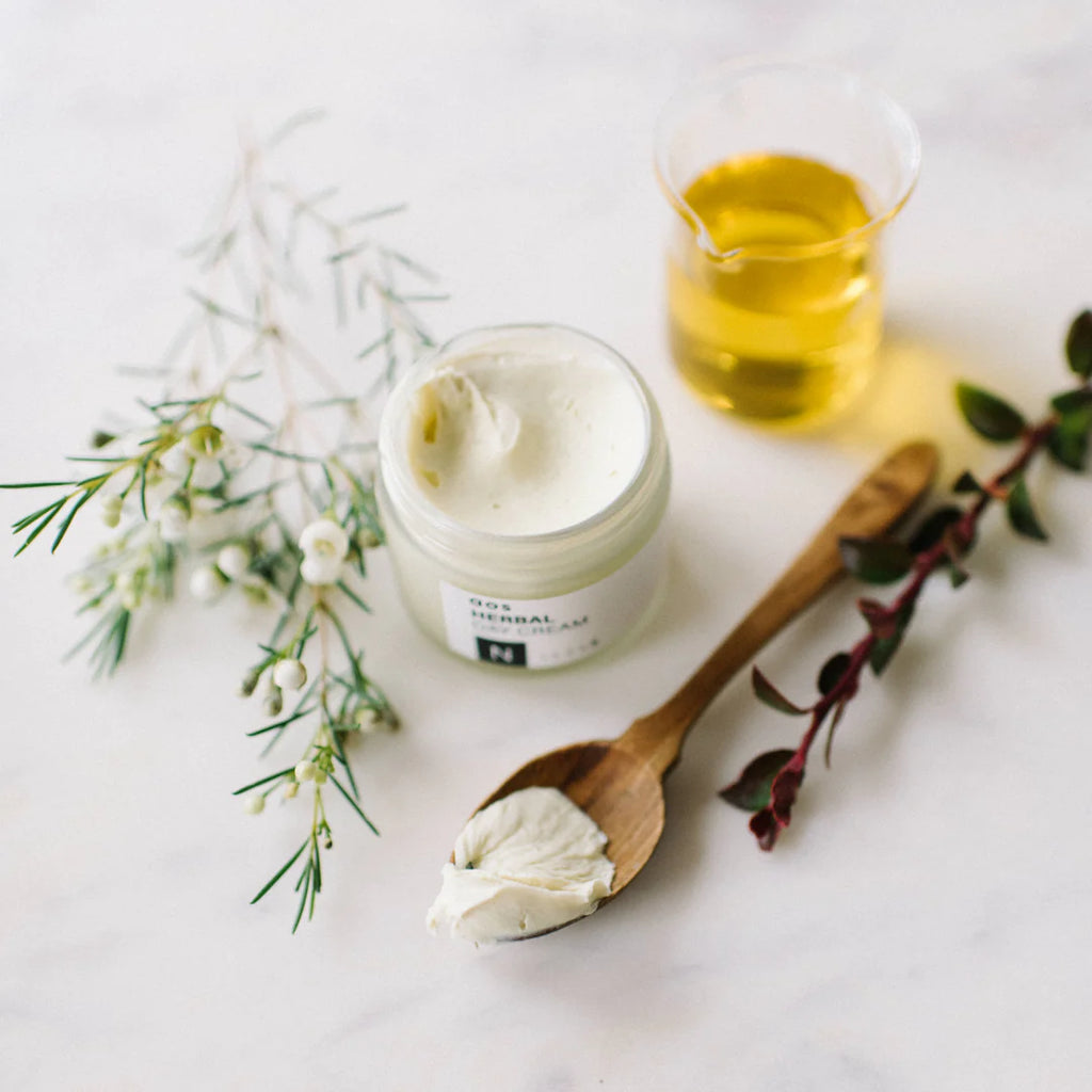 A jar of natural cream with a wooden spoon, a small vase of oil, and sprigs of herbs on a marble surface.