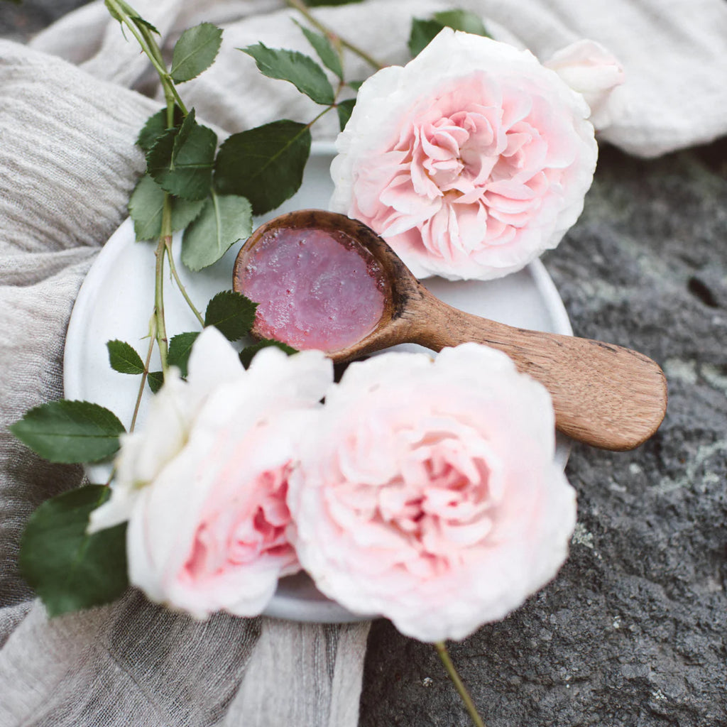 Pink rose petals and a wooden spoon with a skincare product on a white plate, set against a textured gray background.