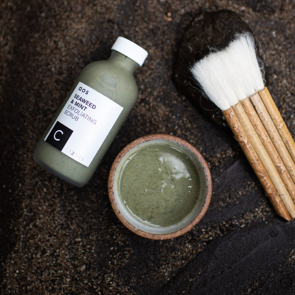 A bowl of seaweed exfoliating scrub with a brush and a bottle of skincare product on a dark surface.