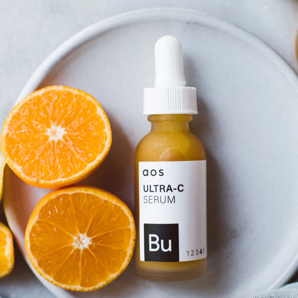 A bottle of ultra-c serum with a dropper on a plate next to sliced oranges.