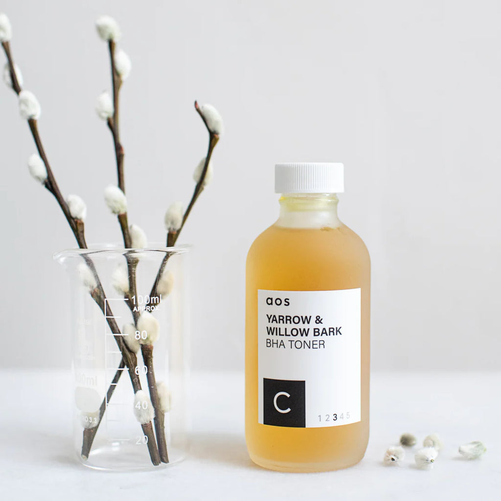 A bottle of yarrow & willow bark bha toner on a plain background with a beaker and pussy willow branches.