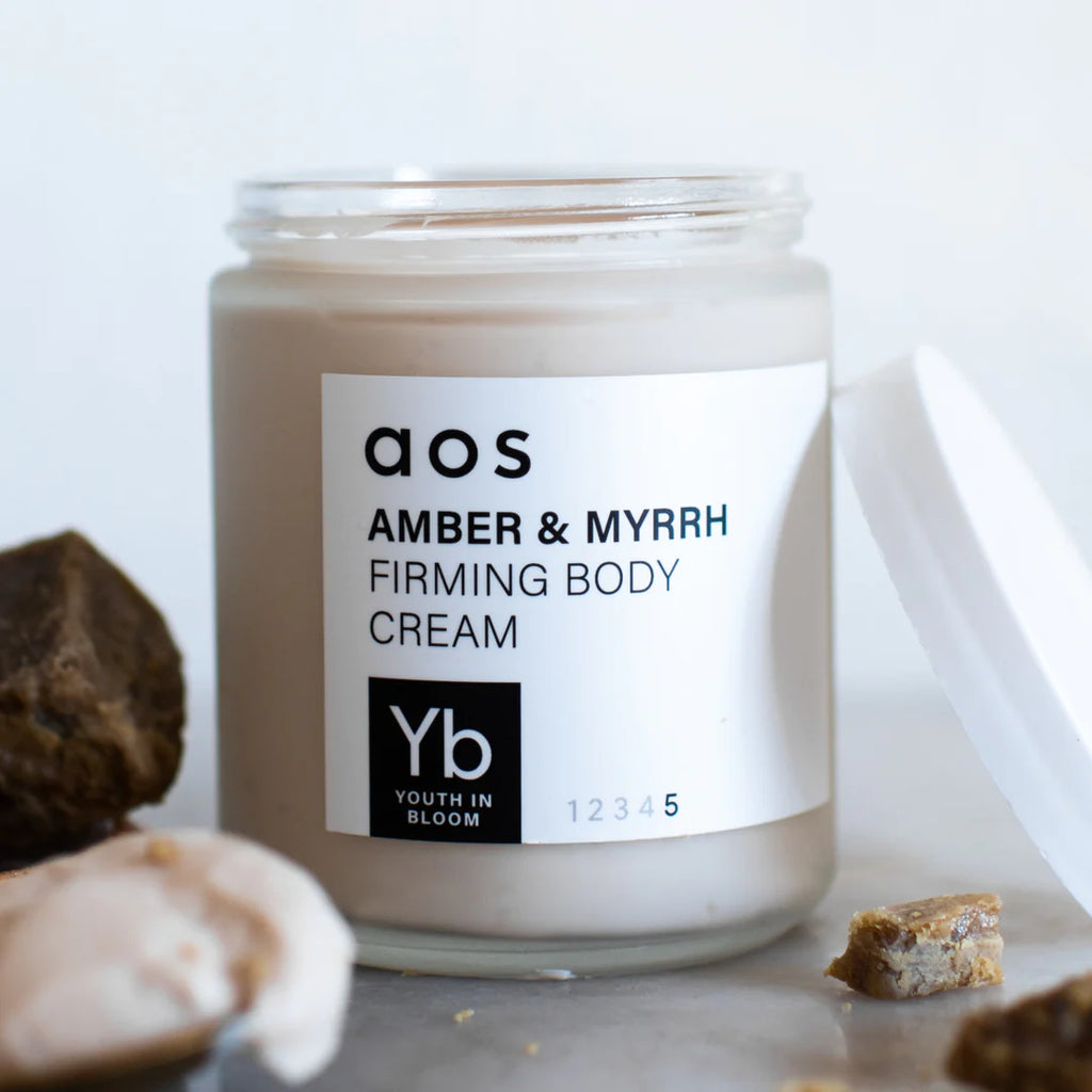 A jar of amber & myrrh firming body cream with an open lid, displayed alongside small stones.
