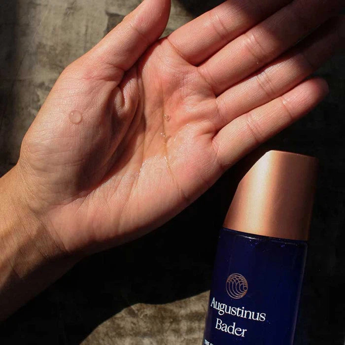 An open hand displaying a few drops of a product next to an augustinus bader bottle.