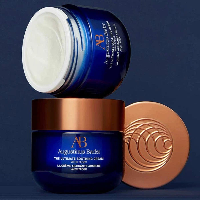 Two jars of augustinus bader moisturizer, one open with cream visible, against a blue background with a metallic cap lying beside them.