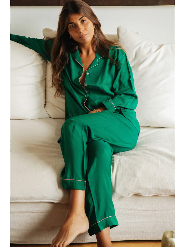 A woman in Germaine des Prés Camille Emerald Pyjamas, made from a blend of green cotton and silk, sits on a white couch with one arm resting on the back.