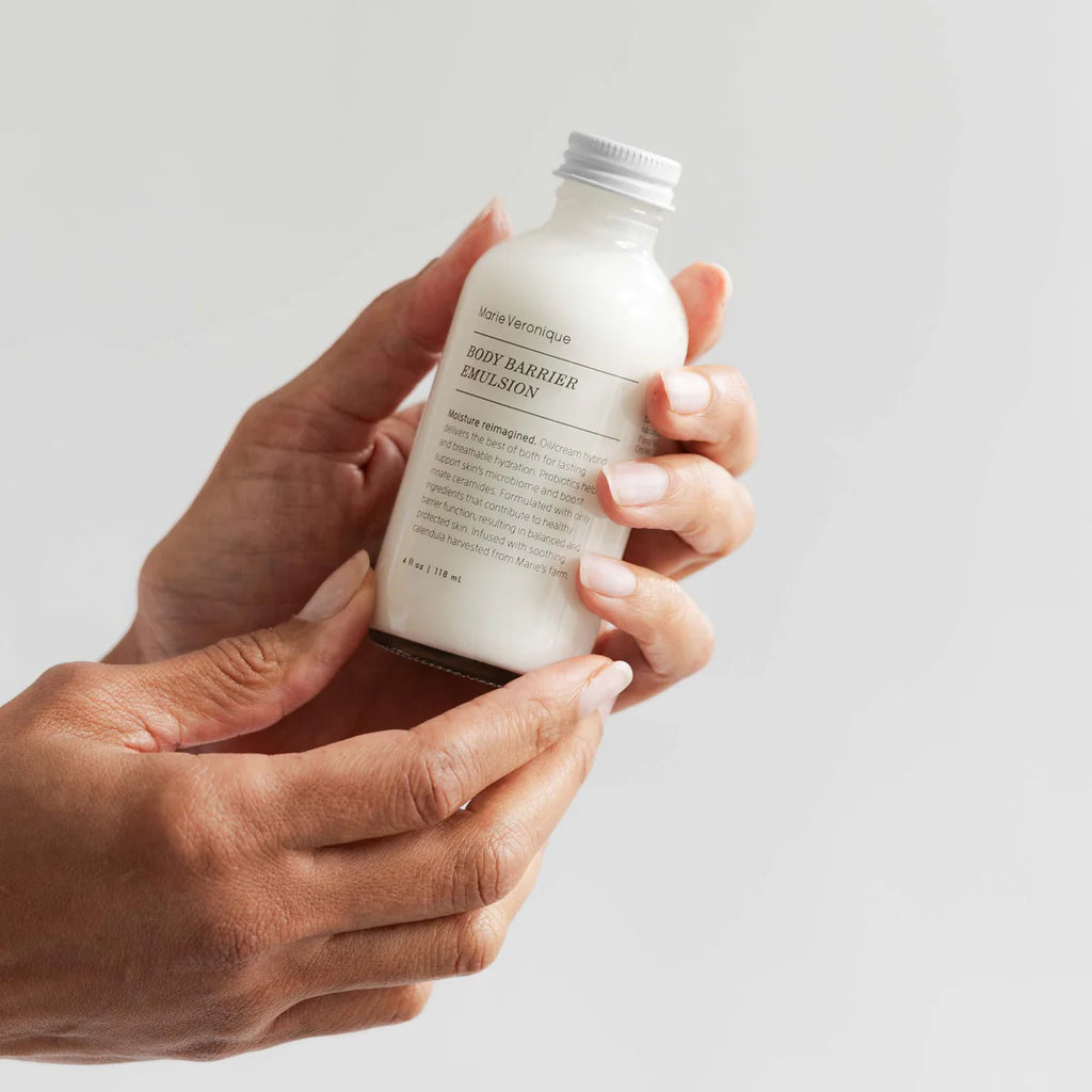 A person holding a bottle of Marie Veronique Body Barrier Emulsion skincare product, enhanced with probiotics, against a light background.