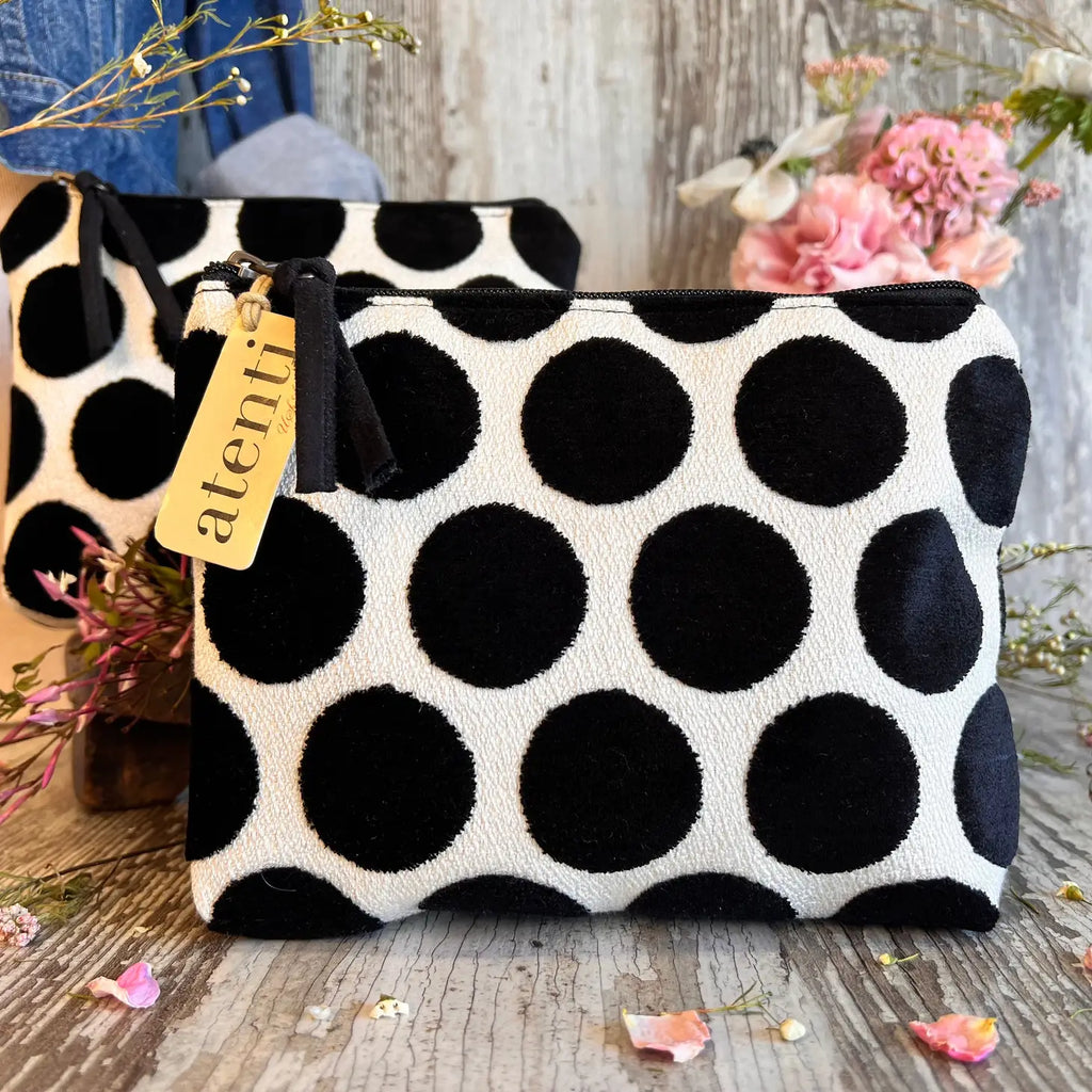 Two black and white polka-dot fabric pouches on a wooden surface with dried flower petals and a pink flower bouquet in the background.