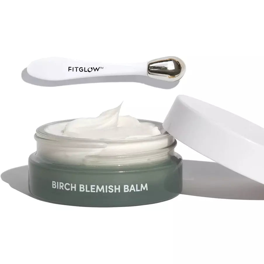 A jar of birch blemish balm with a small application tool and a dollop of cream on a white surface.
